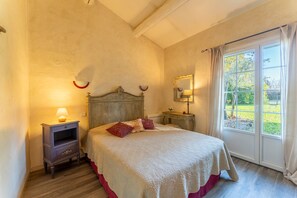 Quarto com cama, mesas de cabeceira, janela com cortinas e vista para o verde lá fora.