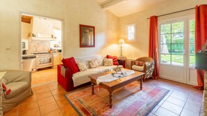 A living room with a sofa, a coffee table, a wicker chair, a kitchen in the background, and a view through a glass door. - Provençal Gîte "La Rivière" – 4 Bedrooms, Private Terrace, Pool & Garden by the Sorgue (Velleron)
