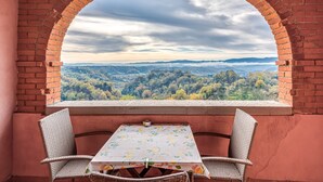 Un balcone con arco in mattoni, sedie in vimini, un tavolo con tovaglia fiorata e vista su un paesaggio boschivo.