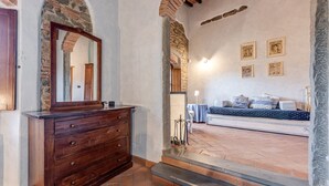 Una stanza con un comò di legno, uno specchio, un letto con cuscini, un muro in pietra e una scala che conduce a un livello superiore.
