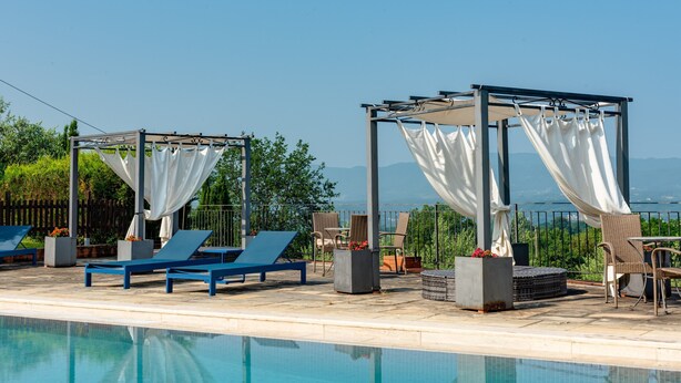 Een zwembadterras met ligbedden, een pergola met witte gordijnen en uitzicht op de bergen.