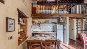 Een eetruimte met een tafel voor vier personen, houten stoelen, een keuken op de achtergrond en een balkon met een hekwerk.