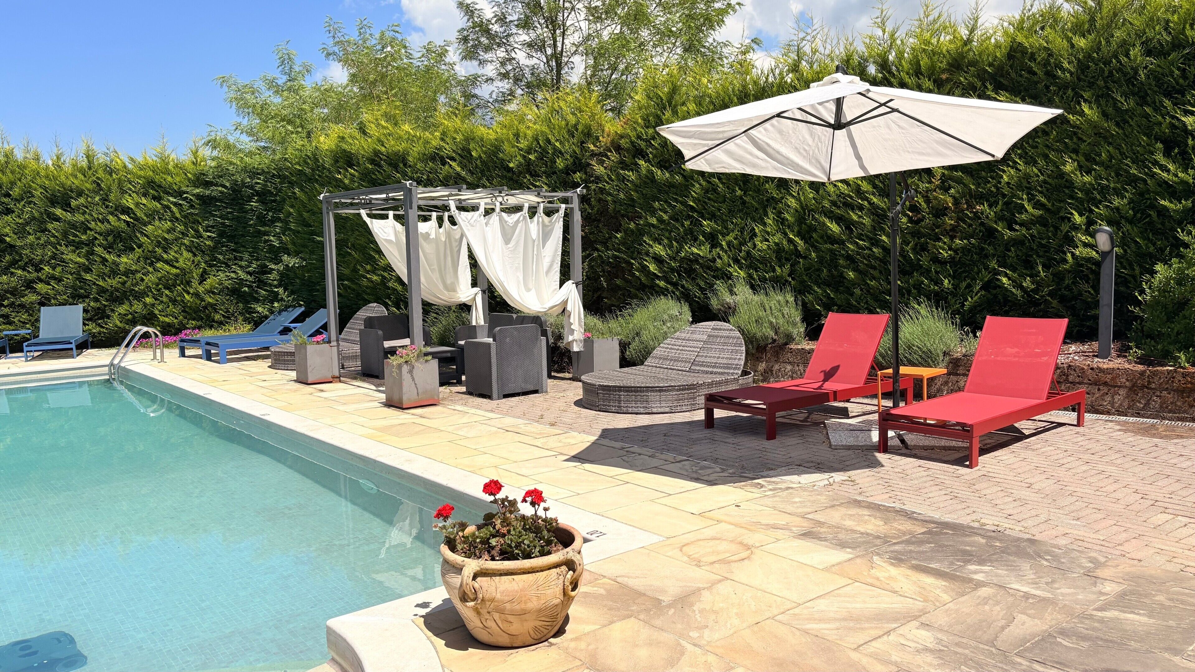 Ein Poolbereich mit Liegestühlen, einer Pergola mit Vorhängen und einem großen Sonnenschirm.