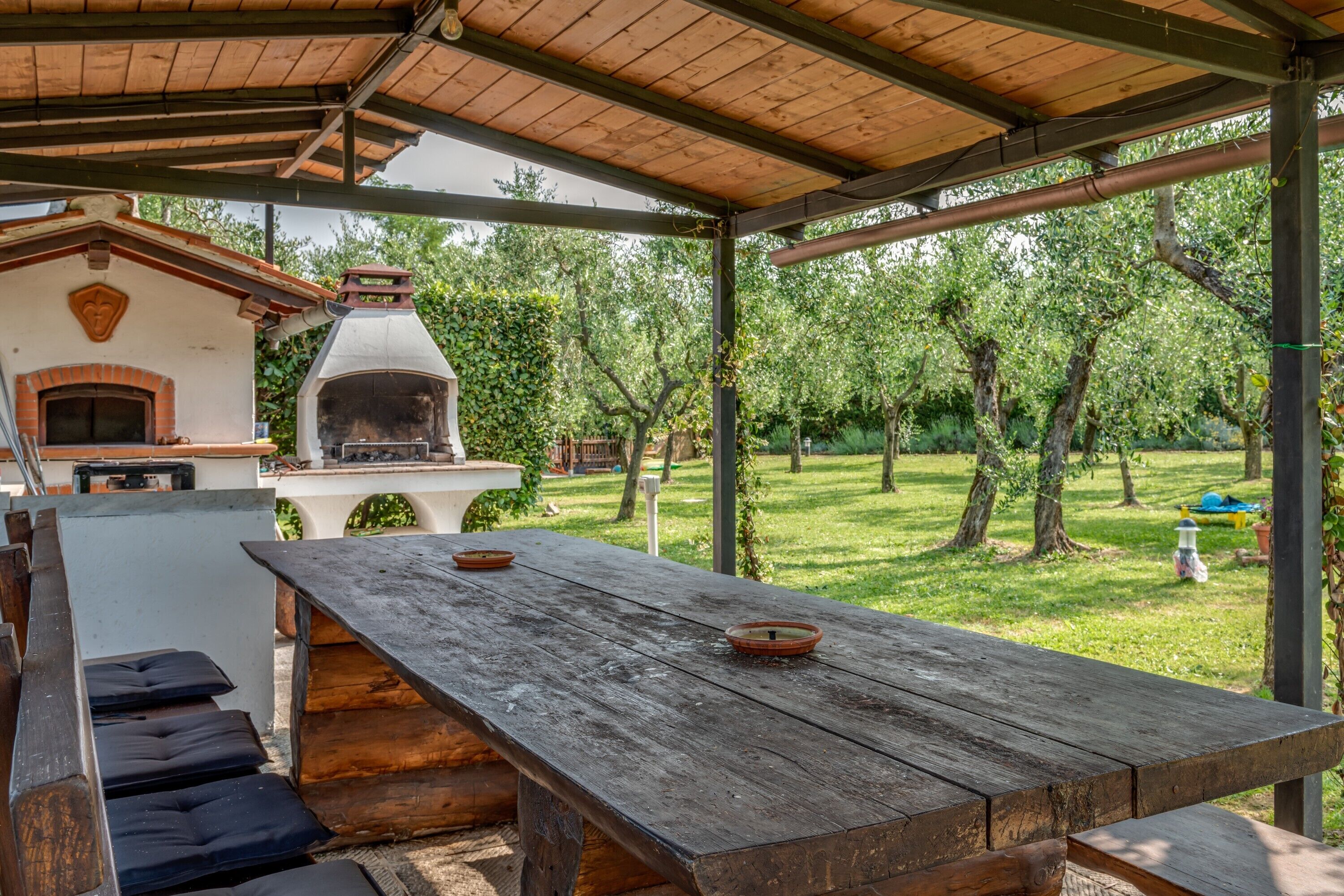 An outdoor patio with a wooden table, a brick oven, and a covered area with a view of a grassy yard and trees.