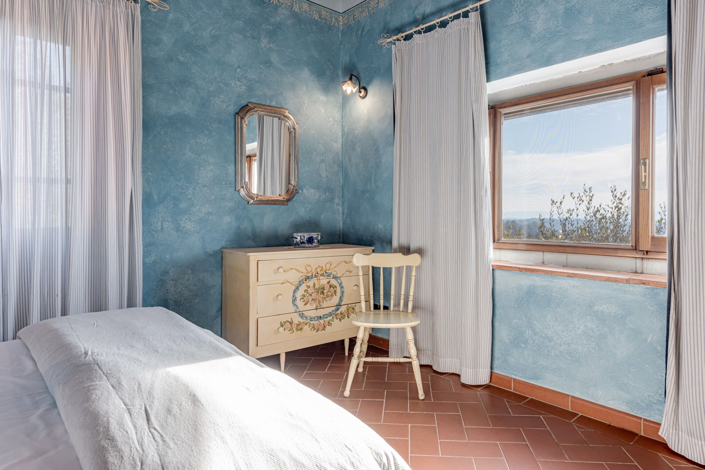 Een slaapkamer met een bed, dressoir, stoel, raam en uitzicht op de omgeving.