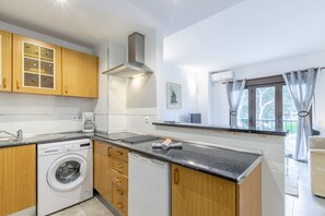 Una cocina compacta con armarios de madera, lavadora integrada, campana extractora de acero inoxidable y una ventana con cortinas.