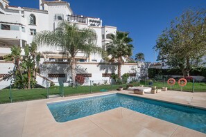A swimming pool with a clear blue water, surrounded by a paved area, palm trees, and a white building with balconies.