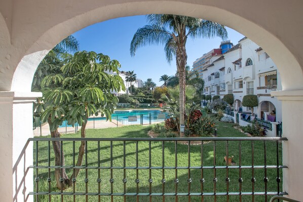A pool area with palm trees, a fenced garden, and a building with balconies and arches.