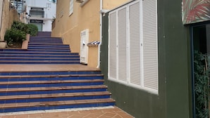 Un edificio residencial con una escalera que conduce a una entrada protegida por una reja, un balcón con barandilla y cielo despejado.