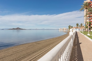 A coastal walkway with a sandy beach, a white railing, and a view of the sea and distant mountains.