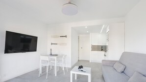 Un salon moderne avec un canapé gris, une table basse blanche, un téléviseur fixé au mur et un coin repas avec des chaises et une table blanches.