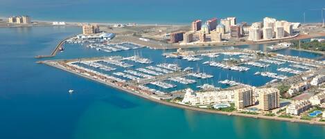An aerial view of a marina with numerous boats, adjacent high-rise buildings, and a clear blue sea.
