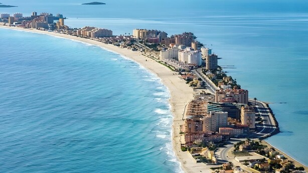 Vista aérea de una ciudad costera con una extensa playa, edificios altos y una cadena montañosa al fondo.
