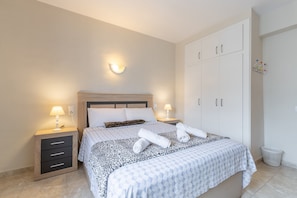 Ein ordentlich bezogenes Bett mit einem gemusterten Überwurf, zwei Nachttischlampen, ein Nachttisch mit Schubladen und ein großer Kleiderschrank.