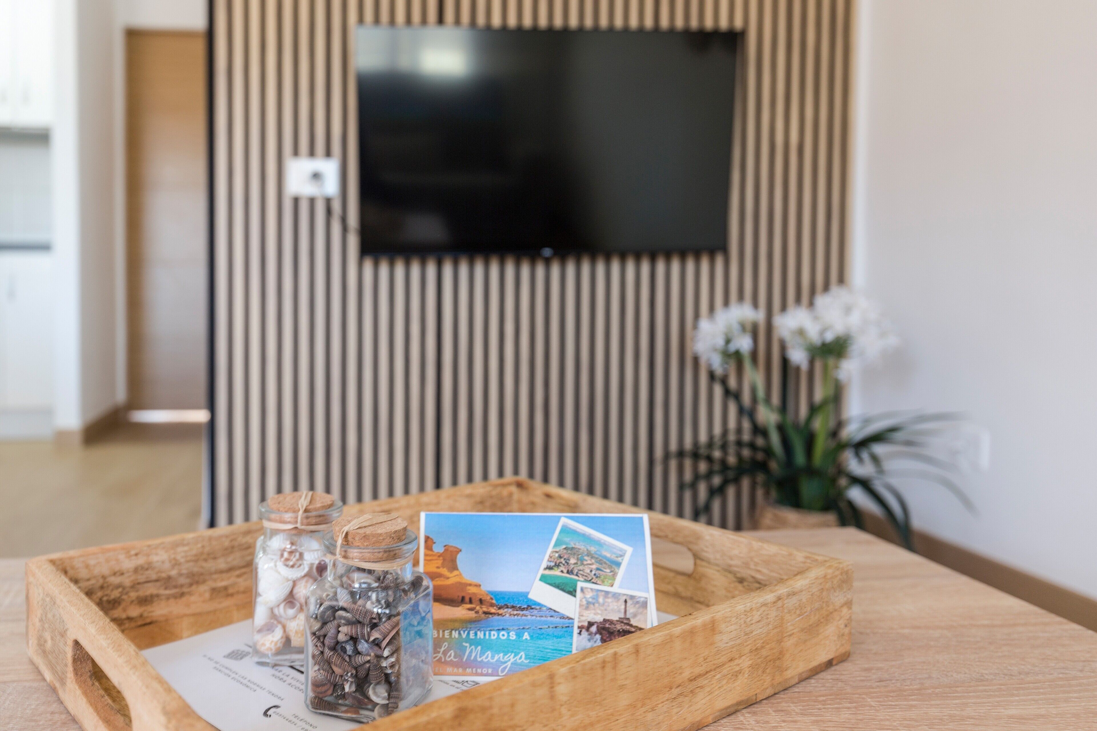 Una bandeja de madera con una tarjeta de bienvenida, un tarro de vidrio con conchas marinas y un tarro de vidrio con monedas sobre una mesa de madera en una habitación con un televisor montado en una pared a rayas.