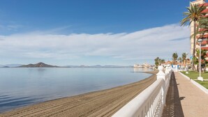 A beach with a white railing, calm water, and distant mountains. - Apartment 'Arena De 2 Mares' with Sea View, Shared Pool and Private Terrace (La Manga)