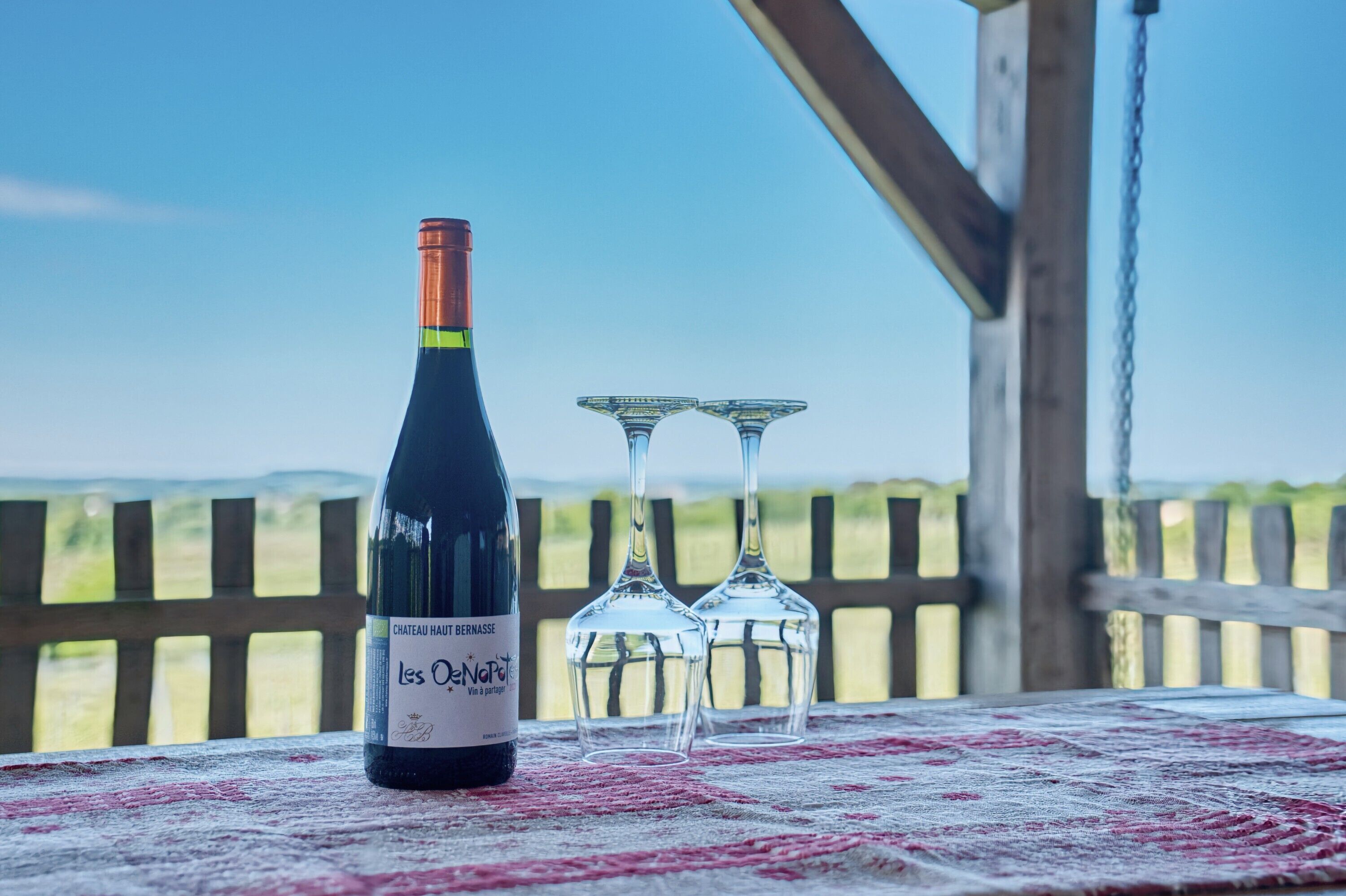 Une bouteille de vin et deux verres sur une table, avec une rambarde en bois et une vue sur la campagne.