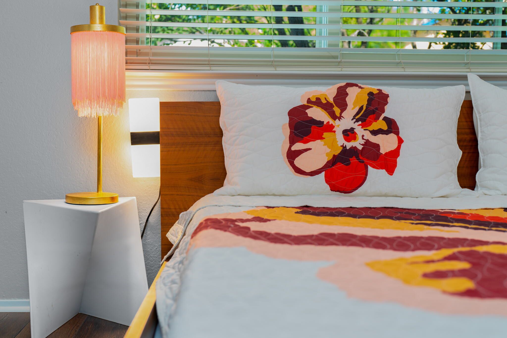A neatly made bed with a colorful floral pillow, a bedside lamp, and a window with blinds.