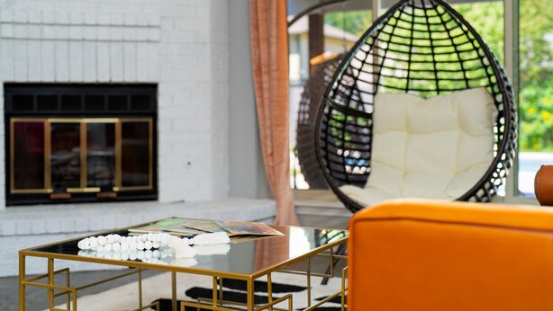 Una habitación con chimenea de ladrillo blanco, una mesa de centro de vidrio con patas doradas y una silla colgante con cojín blanco.