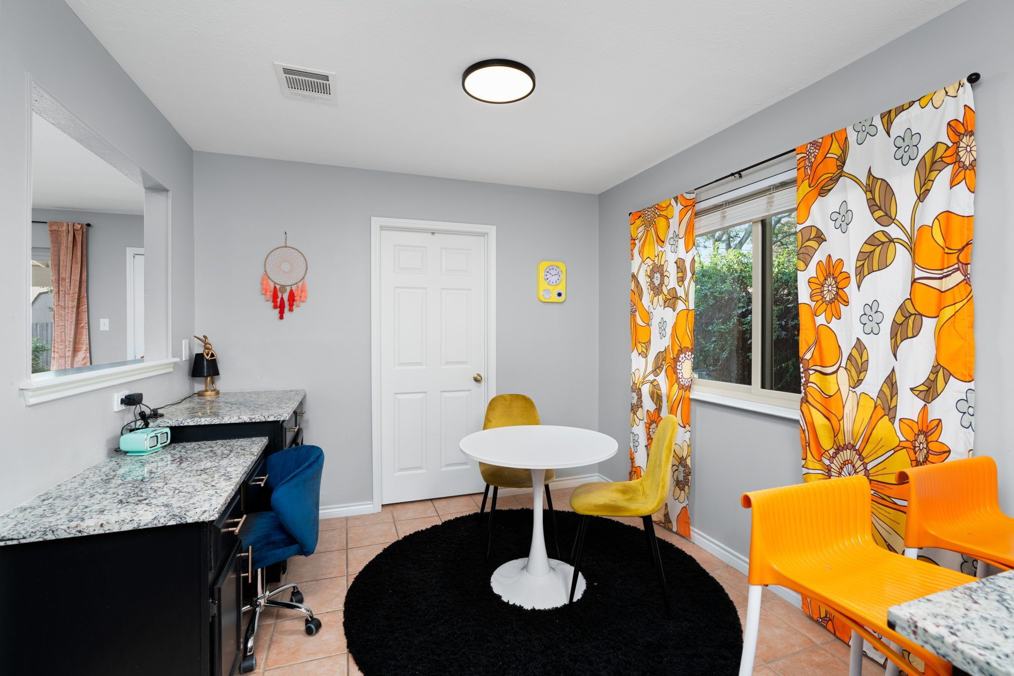 Une pièce de taille modeste avec une porte blanche, une table ronde accompagnée de deux chaises, un petit bureau avec une chaise, un tapis noir et une fenêtre ornée de rideaux à motifs floraux orange.