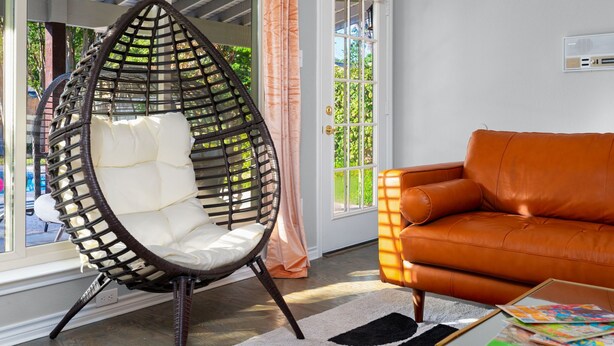 A room with a wicker egg chair, an orange leather sofa, and a window with curtains. - 70s House Fun 6 Bedroom Pool Game Room (Round Rock)