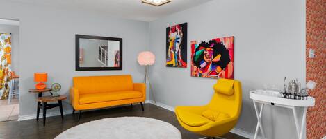 A living room with a round white rug, an orange sofa, a yellow armchair, and a white side table with a lamp and a fan.