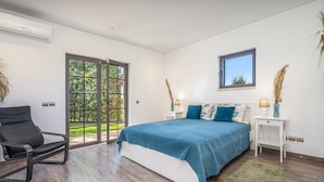 Ein Schlafzimmer mit Bett, Sessel, Nachttisch, Fenster und einer Tür, die auf einen Balkon führt.