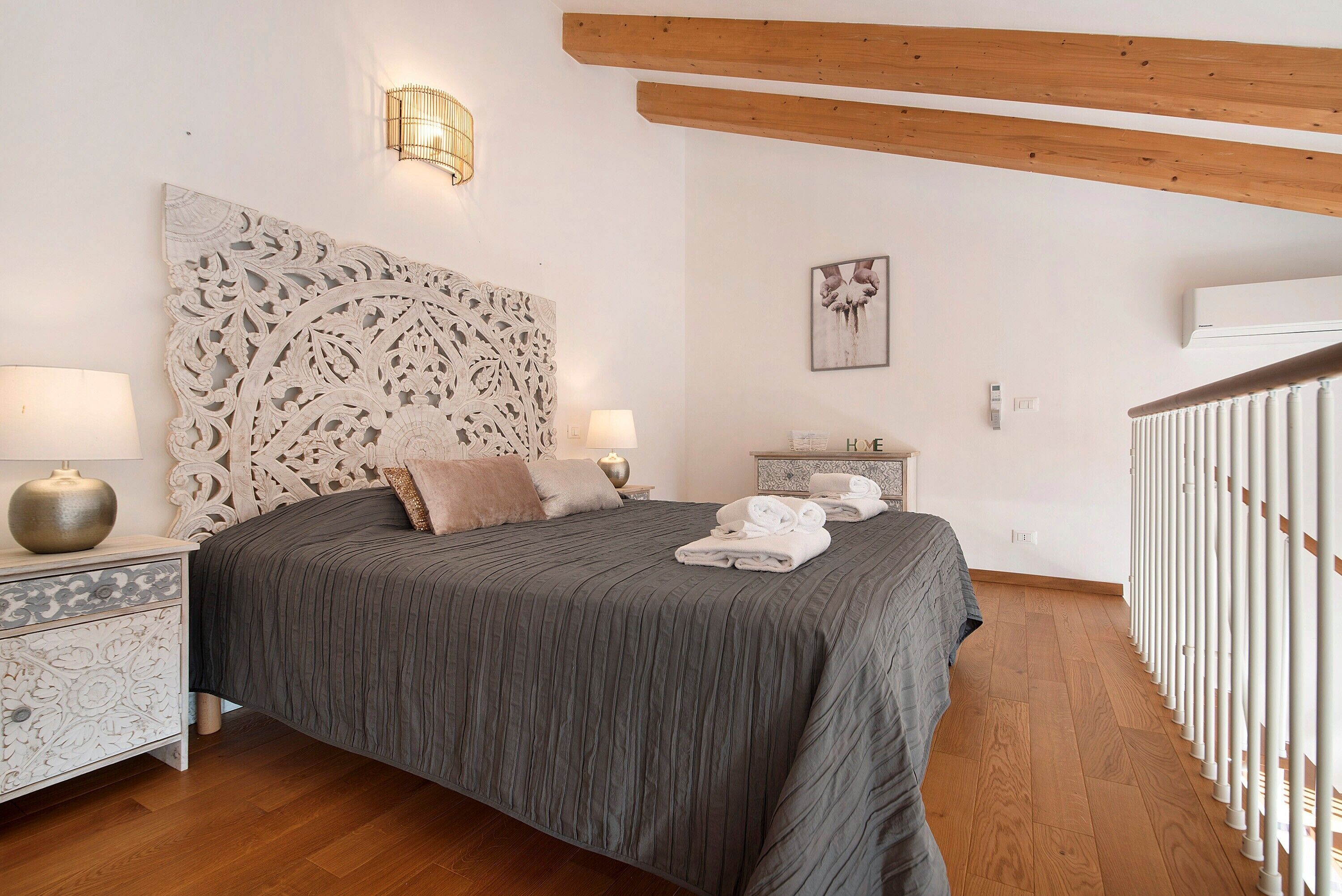 A bedroom with a large bed, two bedside tables with lamps, a decorative headboard, a framed artwork, and a white railing.