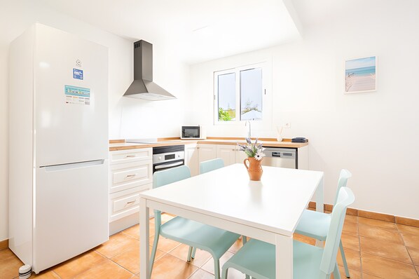 A compact kitchen with a white refrigerator, oven, and dishwasher, a white dining table with chairs, and a window with a view of the outdoors. - Villa 'Las Estrellas Cala Llombards' with Sea View, Wi-Fi and Air Conditioning (Cala Llombards)