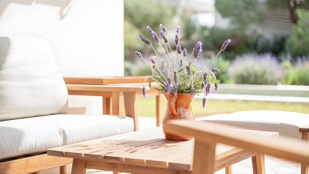 Een houten tafel met een pot lavendel, een witte bank en een houten bankje in een lichte buitenomgeving.