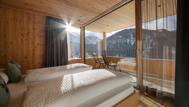 Ein Zimmer mit Holzwänden und -decken, einem Bett mit weißer Bettwäsche, einem Stuhl und einem Balkon mit Bergblick.