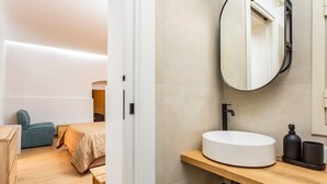 Une chambre d’hôtel moderne avec un plancher en bois, un lit recouvert d’une couette beige, une chaise bleue, un lavabo rond blanc et un miroir au mur.