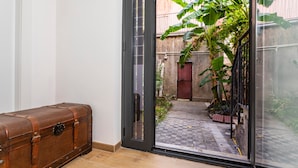 Une pièce dotée d’un plancher en bois, d’une valise en cuir marron et d’une porte-fenêtre coulissante donnant sur un espace extérieur avec un sol à carreaux et des plantes en pot.