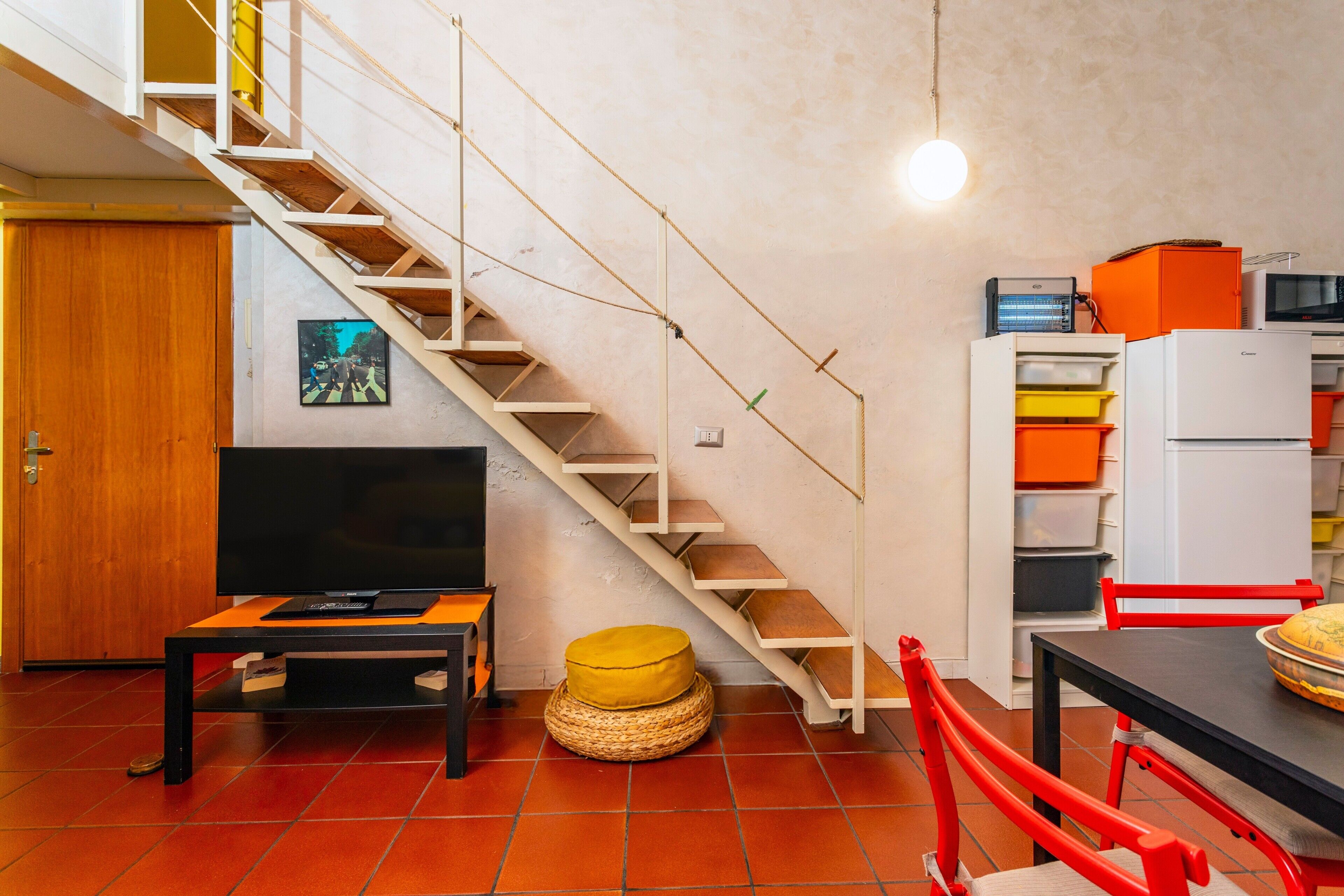 A staircase leading to an upper floor, a television on a stand, a dining table with chairs, and a kitchen area with cabinets and appliances.