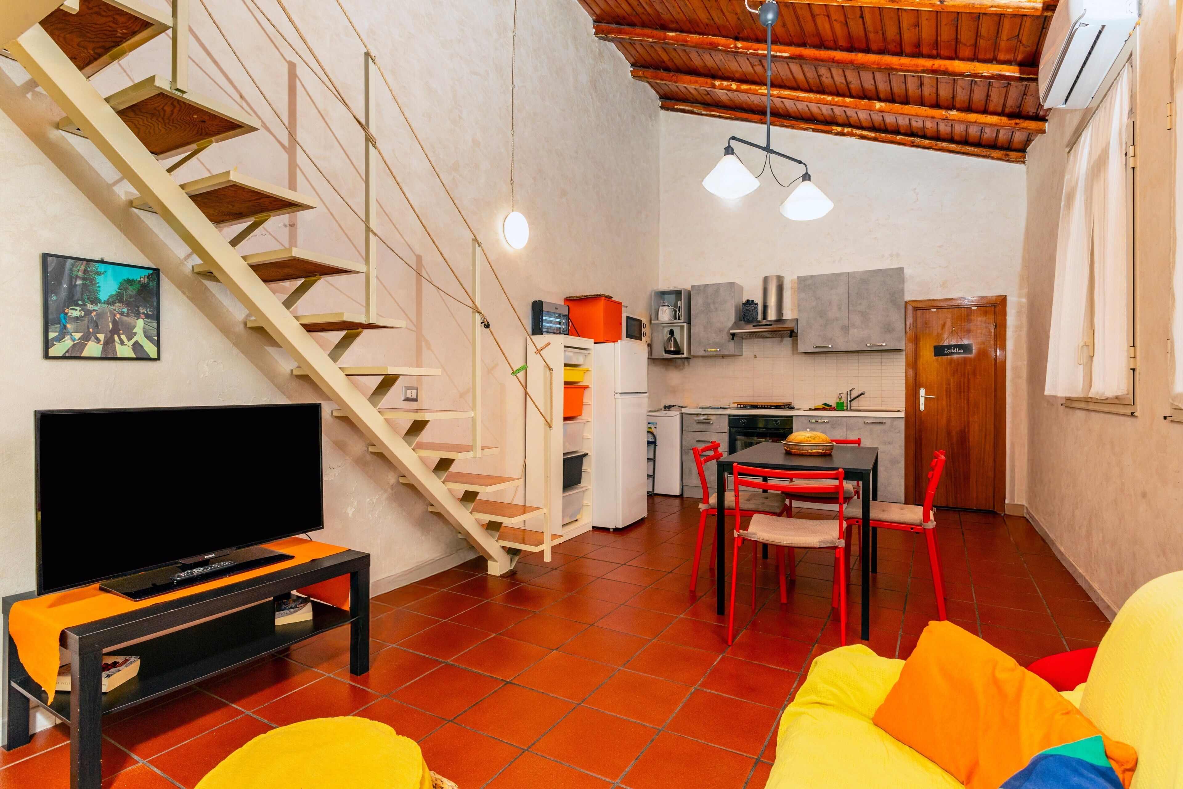 A compact living space with a TV on a stand, a staircase, a dining area with red chairs, and a kitchen area.