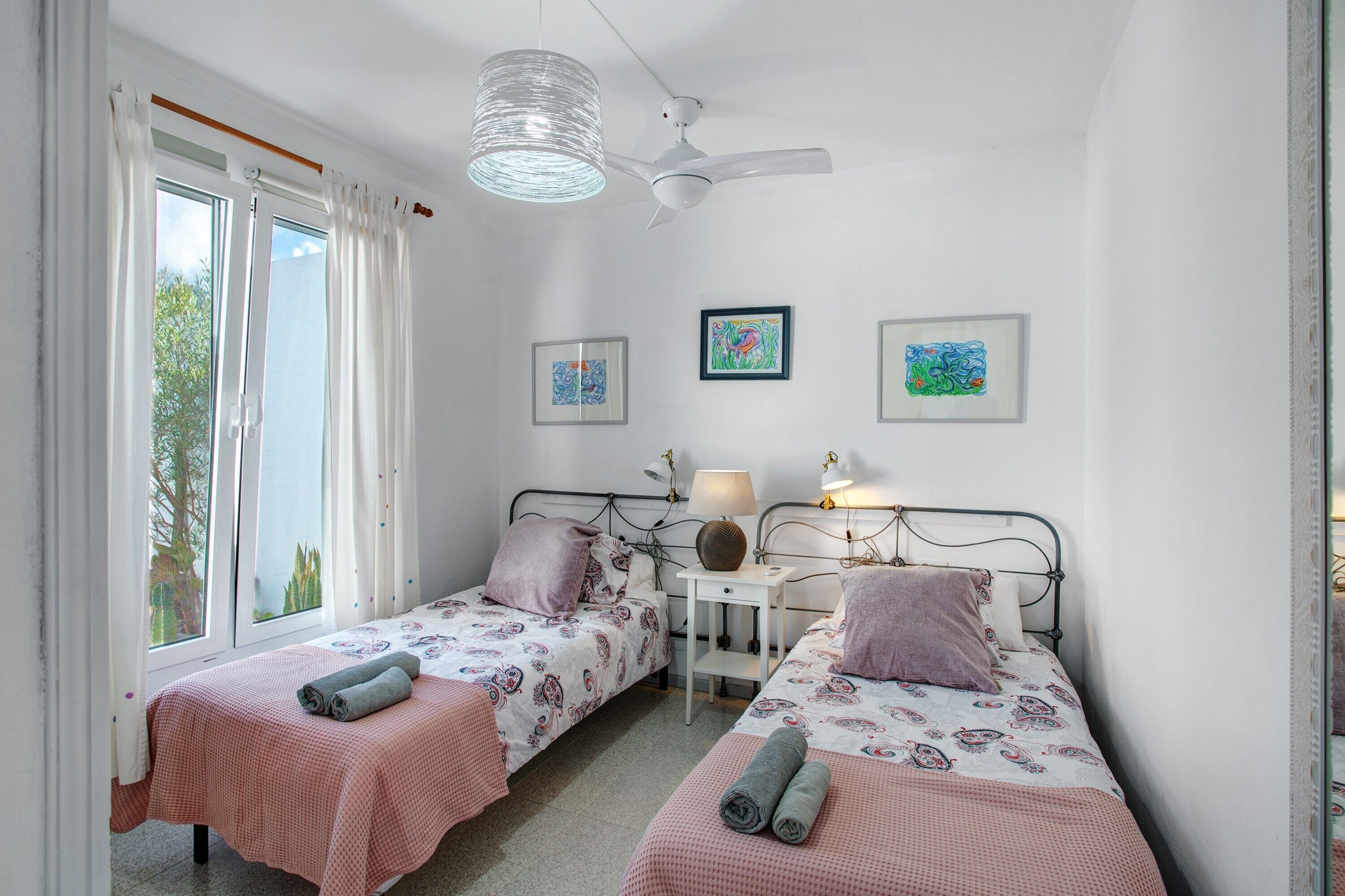 Une chambre avec deux lits, un ventilateur de plafond, une petite table avec une lampe et des œuvres d’art encadrées accrochées au mur.