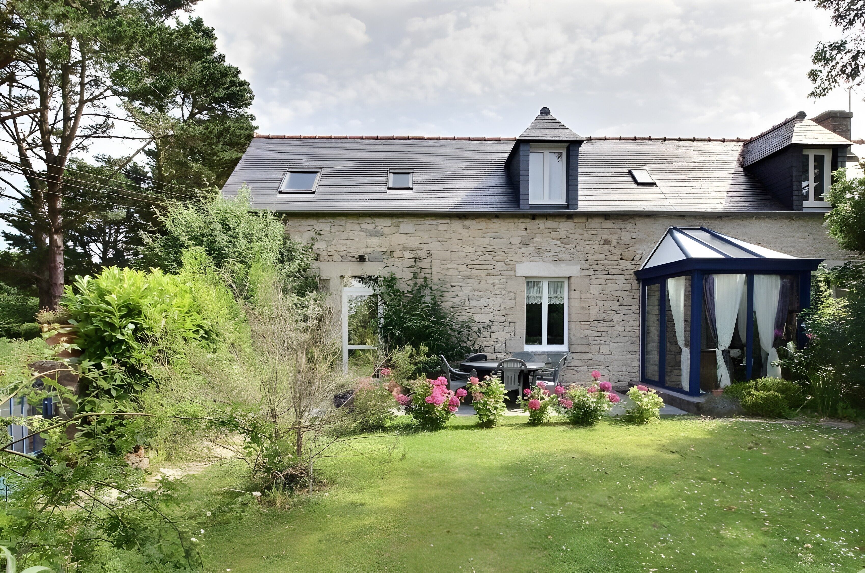 A stone house with a sloped roof, a conservatory, and a well-maintained garden with pink flowers.