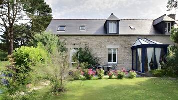 A stone house with a sloped roof, a conservatory, and a well-maintained garden with pink flowers.