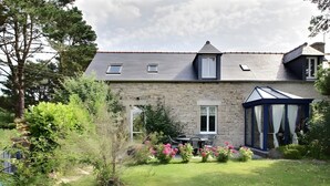 Una casa de piedra con tejado inclinado, un invernadero y un jardín bien cuidado con flores rosadas.