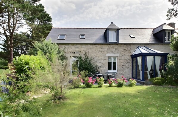 Une maison en pierre avec un toit en pente, une véranda et un jardin bien entretenu agrémenté de fleurs roses.