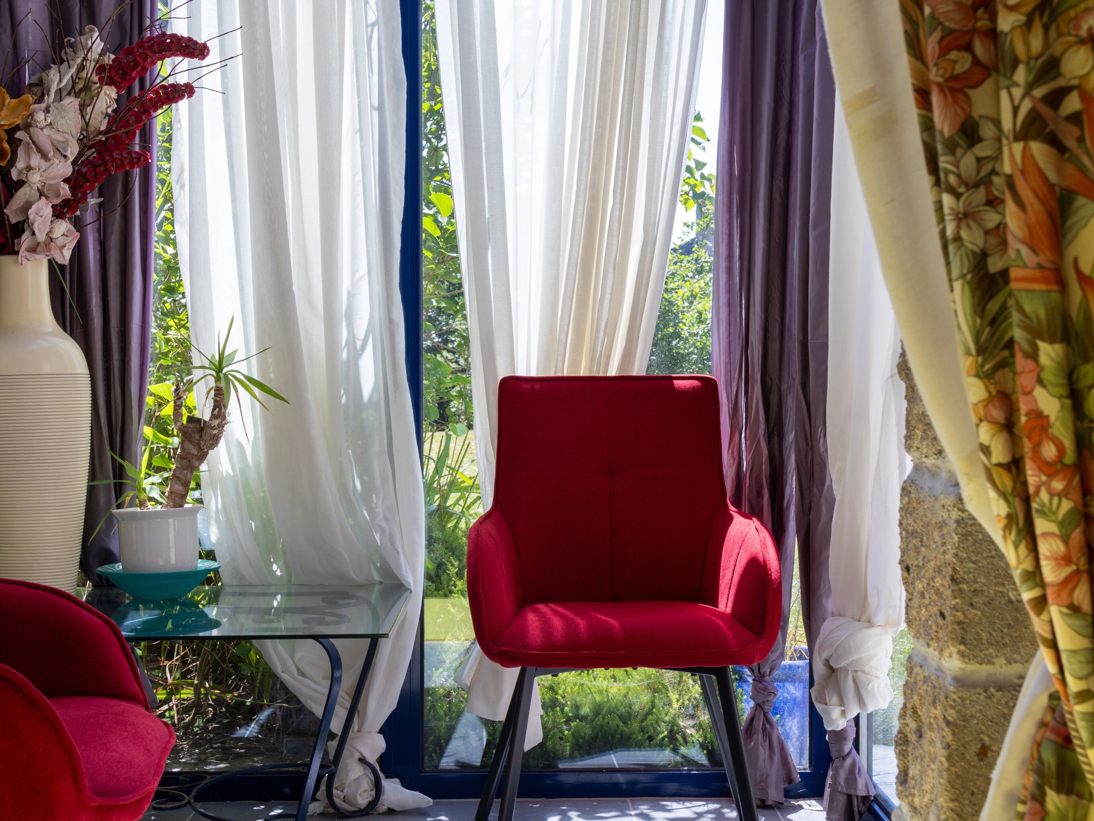 Ein Zimmer mit einem roten Sessel, einem Glastisch, einer weißen Vase mit Blumen und transparenten Vorhängen.