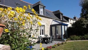 A house with a stone facade, a covered patio with chairs, and a garden with yellow flowers.