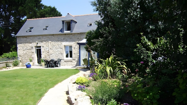 A stone house with a sloped roof, a stone pathway leading to a patio, and a garden with various plants and flowers.