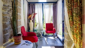 A sunlit room with a stone wall, two red chairs, a small table with a vase of flowers, and a view through a glass door.