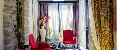 Ein sonnendurchflutetes Zimmer mit einer Steinsäule, zwei roten Stühlen, einem kleinen Tisch mit Blumenvase und Blick durch eine Glastür.