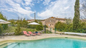 A swimming pool with lounge chairs and umbrellas, surrounded by stone buildings and greenery.