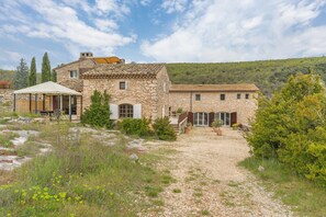 Un bâtiment en pierre avec un chemin de gravier, un espace extérieur abrité avec des sièges, et une vue sur une colline.