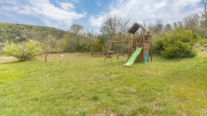 Un parque infantil con un tobogán y columpios en un área verde con árboles y colinas al fondo.