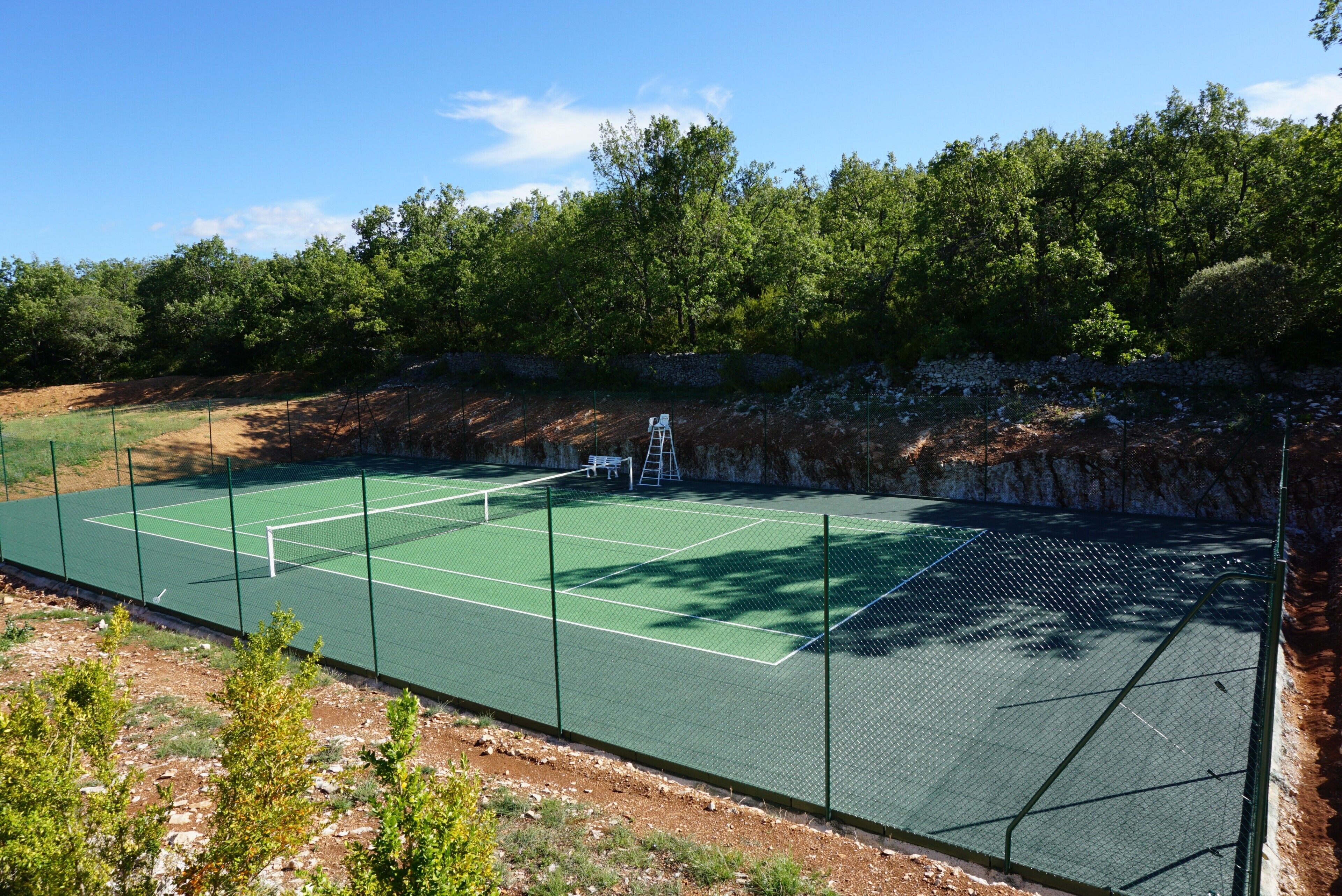 Ein Tennisplatz mit Netz, umgeben von einem Zaun und Bäumen im Hintergrund.