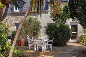 Un bâtiment en pierre doté d’un patio comprenant une table et des chaises, le tout entouré de diverses plantes et arbres.